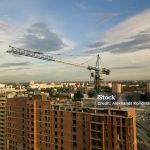 Aerial view of a new modern house under construction with a blue tower crane, red lantern at the end of the crane, building materials on the top floor, bricks, cement, against a blue sky and clouds