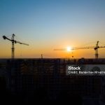 Industrial construction cranes and building silhouettes over sun.