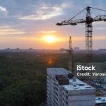 View on industrial landscape with tower cranes and building construction site build skyscrapers at dramatic sunrise sky with green forest on background.