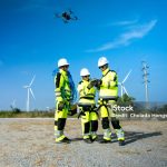 An engineer in a hard hat and safety vest operates a drone to inspect towering wind turbines in a remote area.